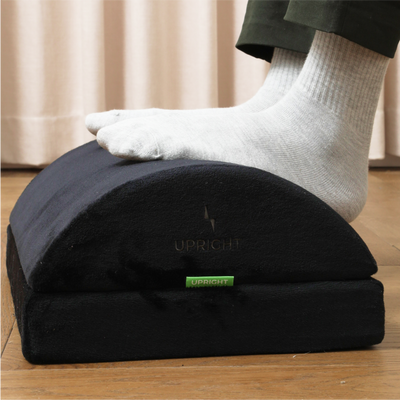 A person's foot resting on a black, memory foam footrest with the brand 'UPRIGHT' visible on the front.